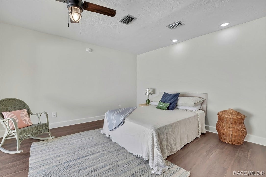 4737 S Sawmill Way, Homosassa, FL 34448 Same bedroom, other angle: bed with white bedding and a striped throw, a navy pillow, white pillows and a green "happy place" pillow, a small nightstand with a green lamp, a wicker rocking chair at the left with a pink pillow, a grey rug, dark wood-look floor, a ceiling fan with a lantern light, recessed lights, and a smoke alarm on the wall.