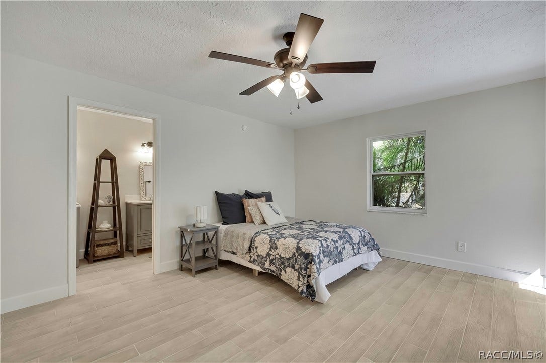 4737 S Sawmill Way, Homosassa, FL 34448 Primary bedroom: bed with navy-and-white floral bedding, dark navy pillows, a patterned pillow and a white bird-print pillow, a grey nightstand with a lamp, a doorway at the left opening to a bathroom vanity area with a dark ladder shelf, a grey vanity and an ornate mirror, a window with palms at the right, light plank tile floor, and a dark ceiling fan with lights.