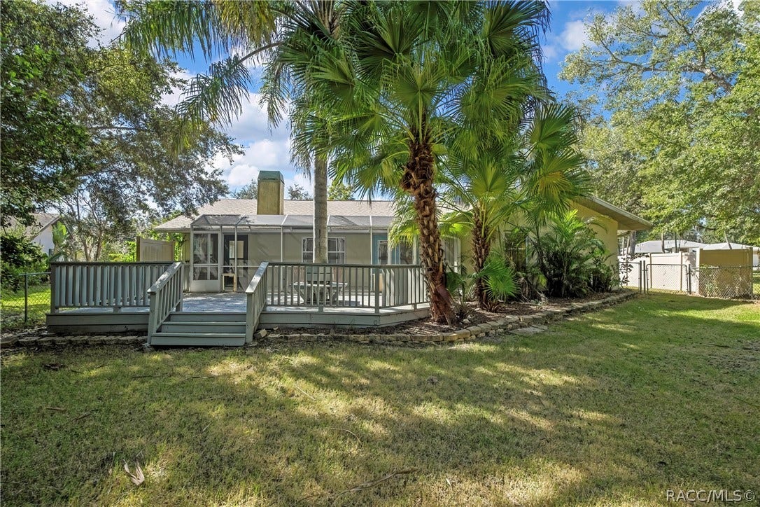 4737 S Sawmill Way, Homosassa, FL 34448 Rear of the house from the lawn: light stucco walls, a shingle roof, a stucco chimney with a metal cap, a screen enclosure with blue doors, a grey wood deck with steps and railing, palm trees, a fenced lawn in the foreground, and neighbouring houses and chain-link and vinyl fences at the sides.