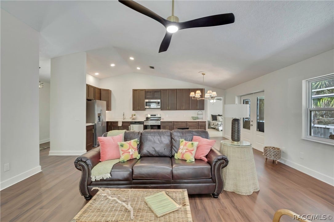 4737 S Sawmill Way, Homosassa, FL 34448 Open living room and kitchen: vaulted ceiling, black ceiling fan with light, white stone fireplace at the right with a wood mantel, mesh screen, black tile hearth and black fireplace tools, brown leather sofa with pink and patterned pillows and a green throw, a wicker trunk coffee table, a beige armchair, dark kitchen cabinets with stainless range, microwave and dishwasher, a sink with a gooseneck faucet, a dining chandelier, French doors to the porch, windows, and wood-look plank floor.