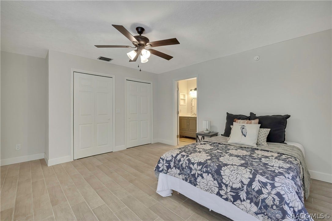 4737 S Sawmill Way, Homosassa, FL 34448 Primary bedroom, third angle: bed with navy-and-white floral bedding and a white bird-print pillow, two white closet doors at the left, a doorway in the centre opening to the bathroom vanity area, a grey nightstand with a lamp, light plank tile floor, white walls, and a dark ceiling fan with lights.
