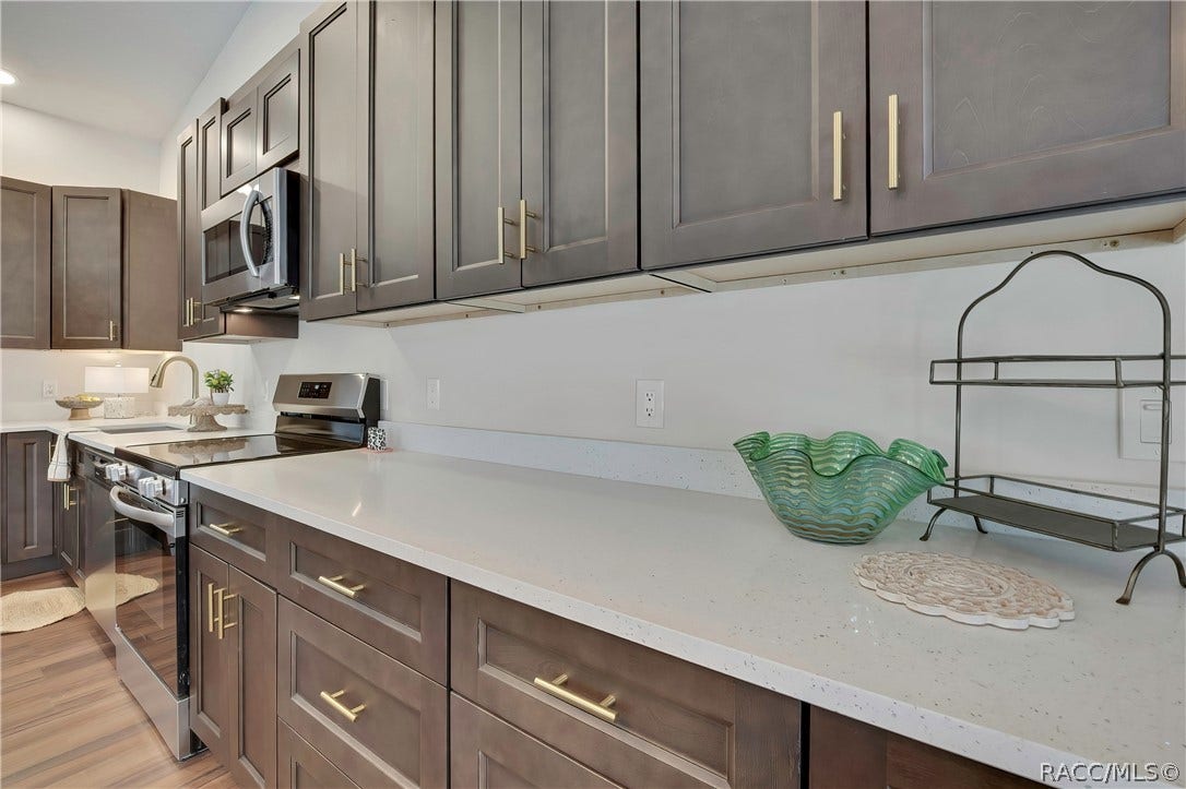 4737 S Sawmill Way, Homosassa, FL 34448 Kitchen counter run: dark cabinets with gold bar pulls, white speckled counters, an electric glass-top range and a microwave above it, a sink with a gooseneck faucet at the left, a green glass bowl, a black wire rack, a white doily placemat and a small patterned container on the counter, and wood-look plank floor.