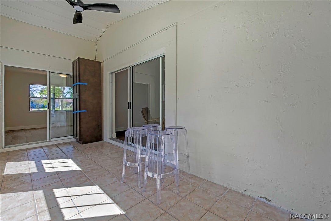 4737 S Sawmill Way, Homosassa, FL 34448 Screened porch, side angle: tiled floor, clear acrylic stools in the centre, sliding glass doors to the bedrooms, a tall dark cabinet with blue painter's tape at the left, a doorway at the left to an empty bedroom, a black ceiling fan at the top of the frame, and a white beadboard ceiling.