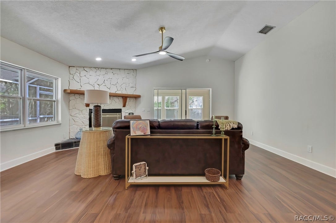 4737 S Sawmill Way, Homosassa, FL 34448 Living room seen from behind the sofa, facing the fireplace: white stone fireplace with a wood mantel, a wicker side table with a tall dark lamp, a gold console table behind the brown leather sofa with a framed picture, a small clock, a basket and green candlesticks, a green throw over the sofa, windows at the left looking onto the screened porch, sliding glass doors in the centre to the Florida room, vaulted ceiling, black ceiling fan with light, recessed lights, and wood-look plank floor.