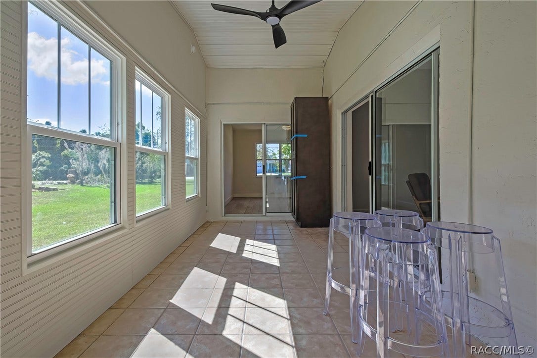 4737 S Sawmill Way, Homosassa, FL 34448 Screened porch, opposite end: tiled floor, windows at the left looking onto the lawn and trees, clear acrylic stools in the foreground at the right, sliding glass doors at the right to the bedrooms, a tall dark cabinet with blue painter's tape in the centre, a doorway at the far end to an empty bedroom, a black ceiling fan at the top of the frame, and a white beadboard ceiling.