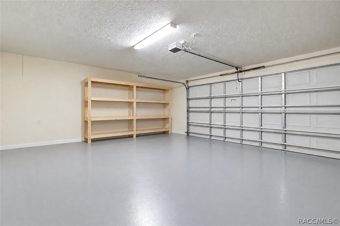 4737 S Sawmill Way, Homosassa, FL 34448 Clean, empty garage interior: smooth grey painted concrete floor, a white sectional garage door with metal rails and a garage door opener, a fluorescent tube light, a textured ceiling, beige walls, and a light wood empty shelving unit with two bays and four shelves against the back wall. No mower, boxes or clutter in this shot this is a different, newer garage photo than the two cluttered garage shots in the old set.