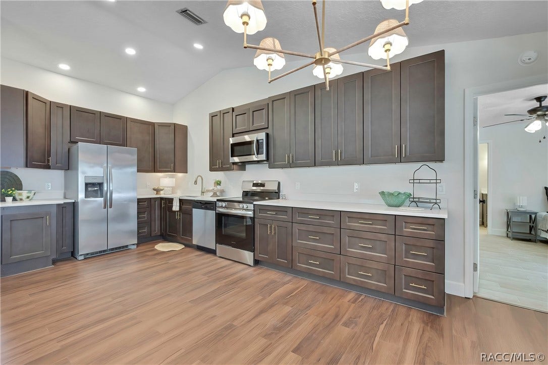 4737 S Sawmill Way, Homosassa, FL 34448 Wide kitchen view: dark grey-brown cabinets with gold bar pulls, white speckled counters, a stainless side-by-side refrigerator with a water/ice dispenser, an electric glass-top range and oven, a microwave above the range, a dishwasher, a sink with a gooseneck faucet and a towel, a small plant, a green glass bowl and a black wire rack on the counter, a chandelier at the top of the frame, recessed lights, wood-look plank floor, and a doorway at the right opening to the primary bedroom