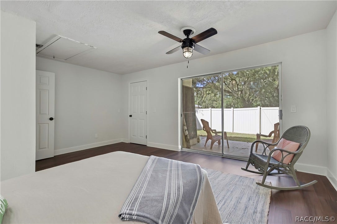 4737 S Sawmill Way, Homosassa, FL 34448 Same bedroom, third angle: bed in the foreground with white bedding, a striped throw and a green pillow at the edge, a wicker rocking chair at the right with a pink pillow, a grey rug, dark wood-look floor, sliding glass doors in the centre to a fenced patio with a white vinyl fence and lawn chairs outside, white doors at the left, an attic hatch in the ceiling, and a ceiling fan with a lantern light.