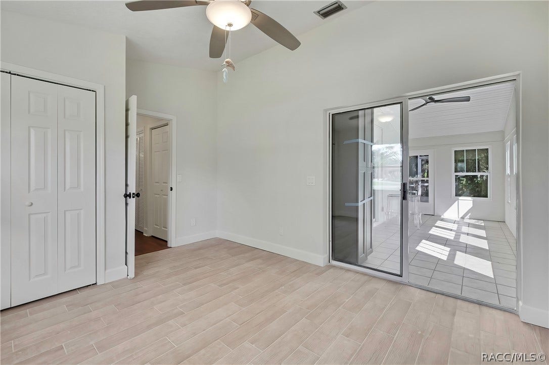 4737 S Sawmill Way, Homosassa, FL 34448 Empty bedroom: light plank tile floor, white double closet doors at the left, an open doorway to a hallway with a dark wood floor, a sliding glass door at the right to the screened porch with a tiled porch floor and chairs visible through it, a ceiling fan with a dome light, and white walls. No furniture.