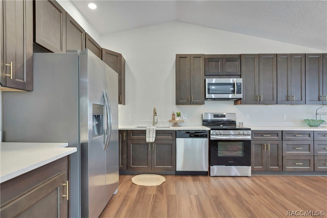 4737 S Sawmill Way, Homosassa, FL 34448 Wide kitchen view: dark grey-brown cabinets with gold bar pulls, white speckled counters, a stainless side-by-side refrigerator with a water/ice dispenser, an electric glass-top range and oven, a microwave above the range, a dishwasher, a sink with a gooseneck faucet and a towel, a small plant, a green glass bowl and a black wire rack on the counter, a chandelier at the top of the frame, recessed lights, wood-look plank floor, and a doorway at the right opening to the primary bedroom.