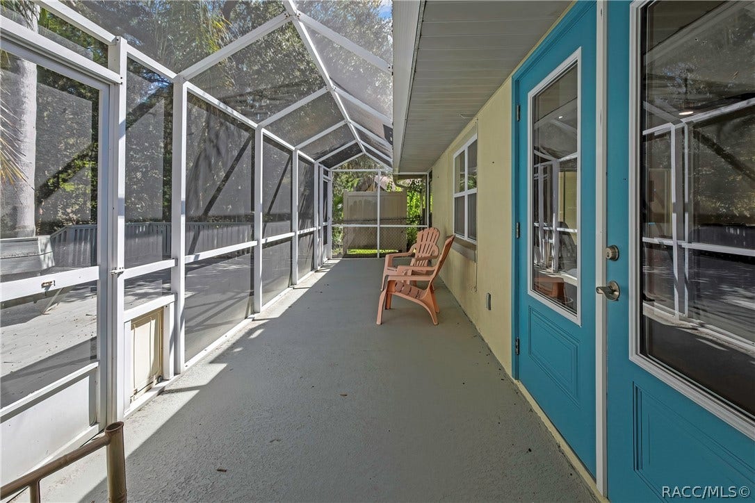4737 S Sawmill Way, Homosassa, FL 34448 Inside the screen enclosure, along the house wall: concrete floor, white aluminium frame and screen roof, blue entry doors with glass at the right, a window, beige plastic chairs in the centre, a screen door with a pet door at the left, and a grey wood deck beyond.