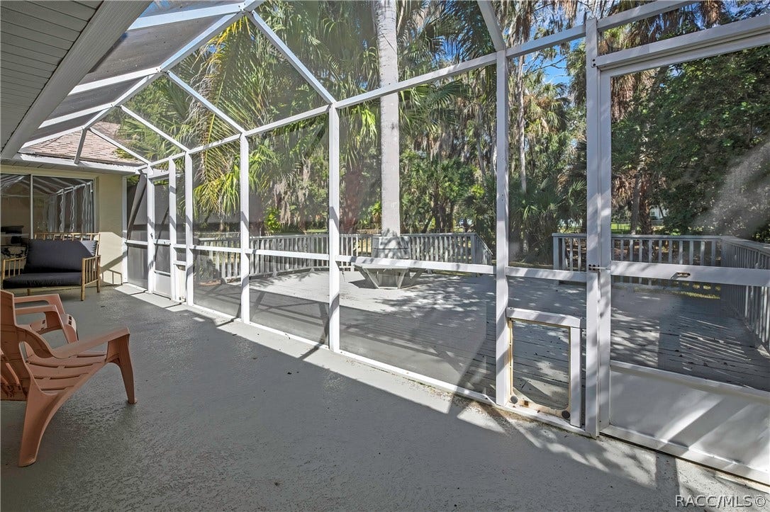 4737 S Sawmill Way, Homosassa, FL 34448 Wood deck: grey wood deck planks, railing, a built-in bench around a palm tree trunk in the centre, steps at the right down to the lawn, the screen enclosure at the left with blue doors, palm trees, a chain-link fence, and lawn and trees beyond.