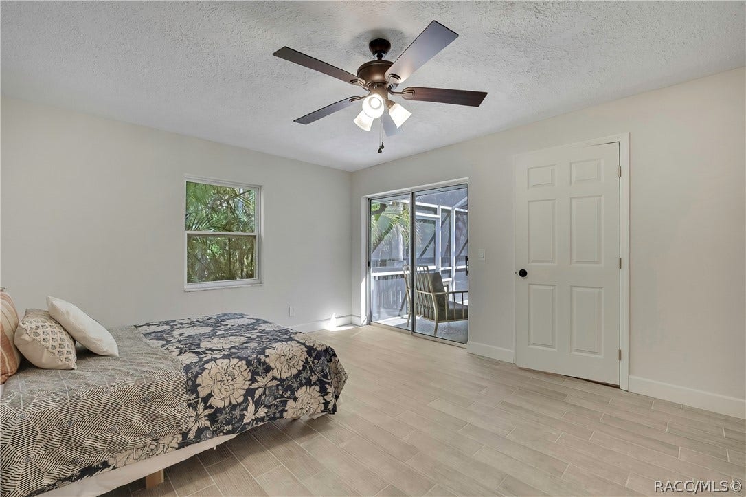4737 S Sawmill Way, Homosassa, FL 34448 Primary bedroom from the porch side: bed with navy-and-white floral bedding, white and patterned pillows, light plank tile floor, a dark 5-blade ceiling fan with lights, a window with palms outside, a sliding glass door to the screened porch with a chair outside, a white closet door at the right, white walls and a textured ceiling.