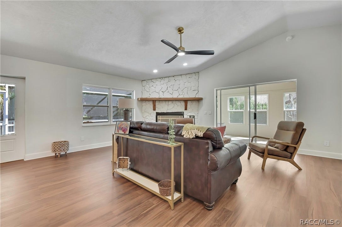 4737 S Sawmill Way, Homosassa, FL 34448 Living room seen from behind the brown leather sofa: vaulted white ceiling, black 3-blade ceiling fan with brass mount and light, recessed lights, white stone fireplace with a wood mantel and black tile hearth, sliding glass doors to the screened porch / Florida room at the right, windows looking onto the porch, a gold console table behind the sofa with a framed picture, glass candlesticks, a small clock and a basket below it, a beige armchair, a green throw over the sofa, wood-look plank floor
