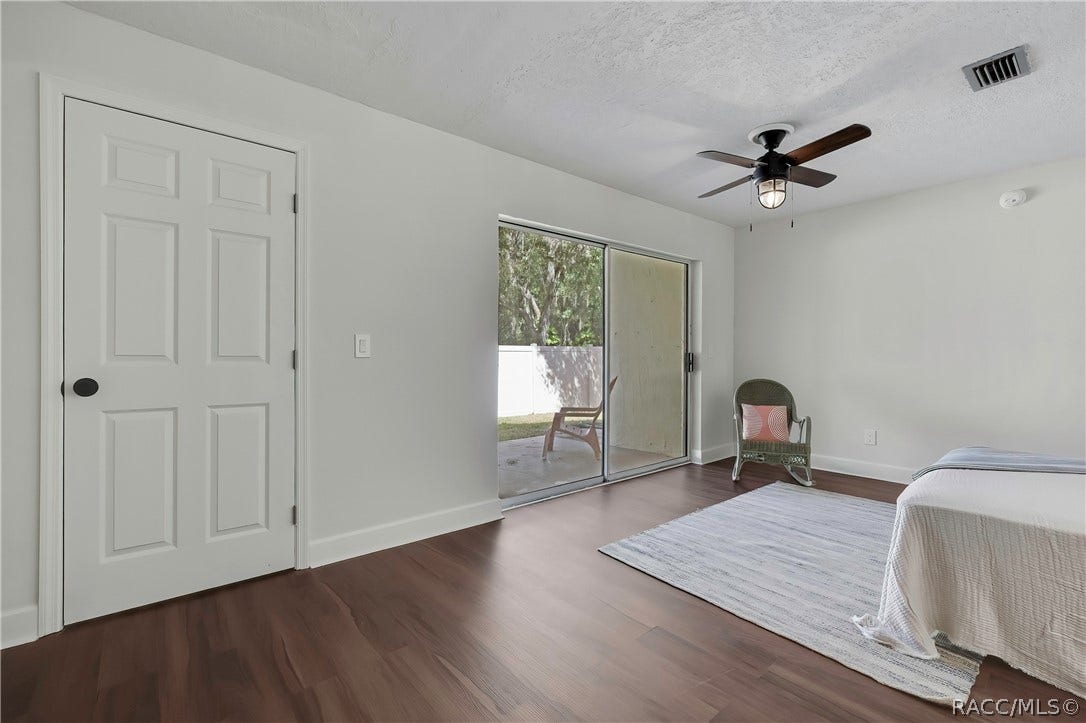 4737 S Sawmill Way, Homosassa, FL 34448 Bedroom: bed at the right with white bedding and a striped throw, a wicker rocking chair with a pink pillow, a grey rug, dark wood-look floor, a ceiling fan with a lantern light, a white door at the left, and a sliding glass door in the centre to a fenced patio with lawn chairs outside and a white vinyl fence, with trees beyond.