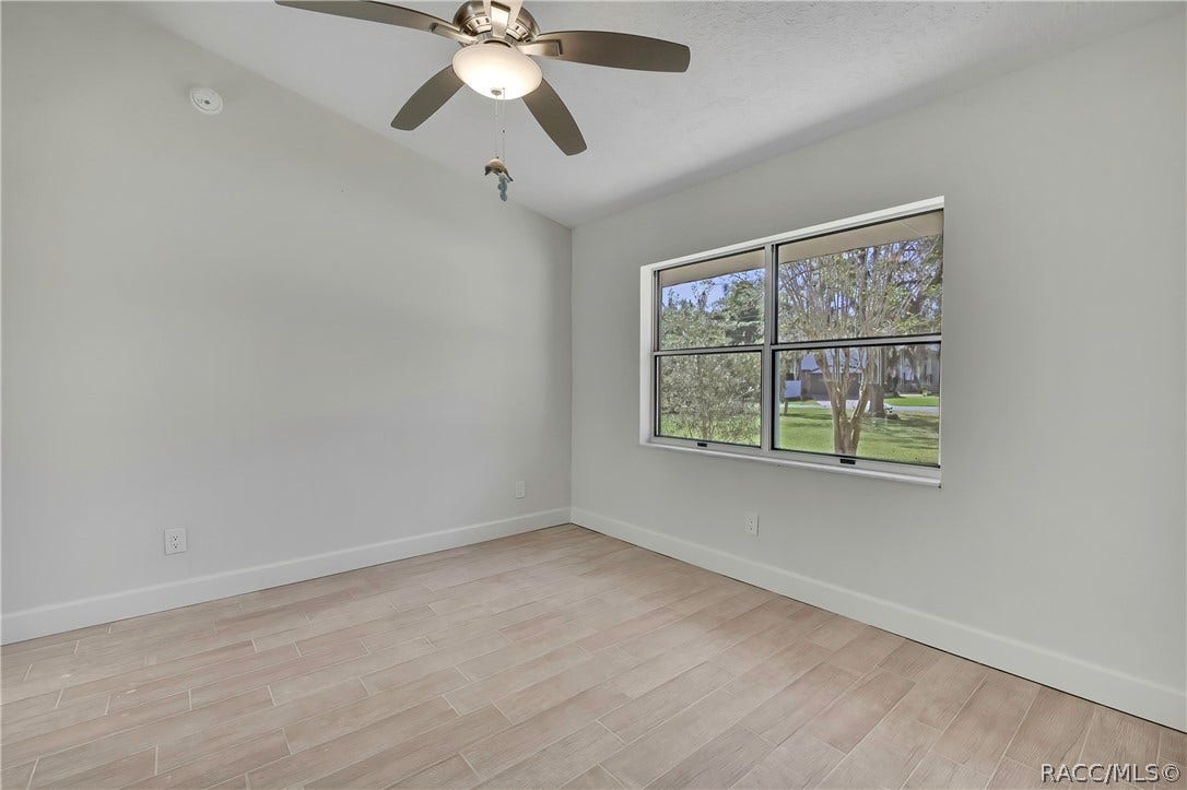 4737 S Sawmill Way, Homosassa, FL 34448 Empty bedroom: light plank tile floor, a large double window looking onto trees, a neighbouring house and lawn, a ceiling fan with a dome light, white walls, wall outlets and a smoke alarm. No furniture.
