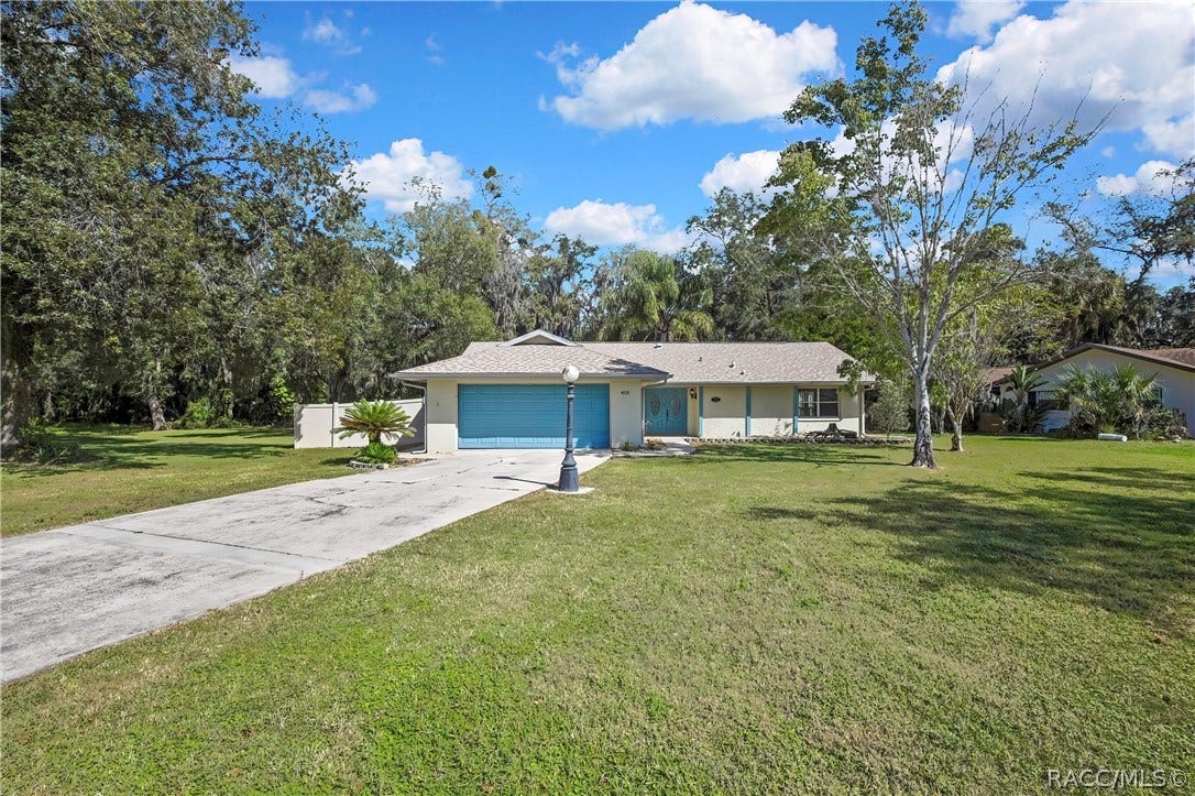 4737 S Sawmill Way, Homosassa, FL 34448 Front of the house seen from the end of the long concrete driveway: blue garage door and blue entry doors, white vinyl fence and a palm plant at the left, a black lamp post with a white globe in the centre of the lawn, green lawn, a neighbouring house at the right, trees and palms behind the house, blue sky with white clouds.
