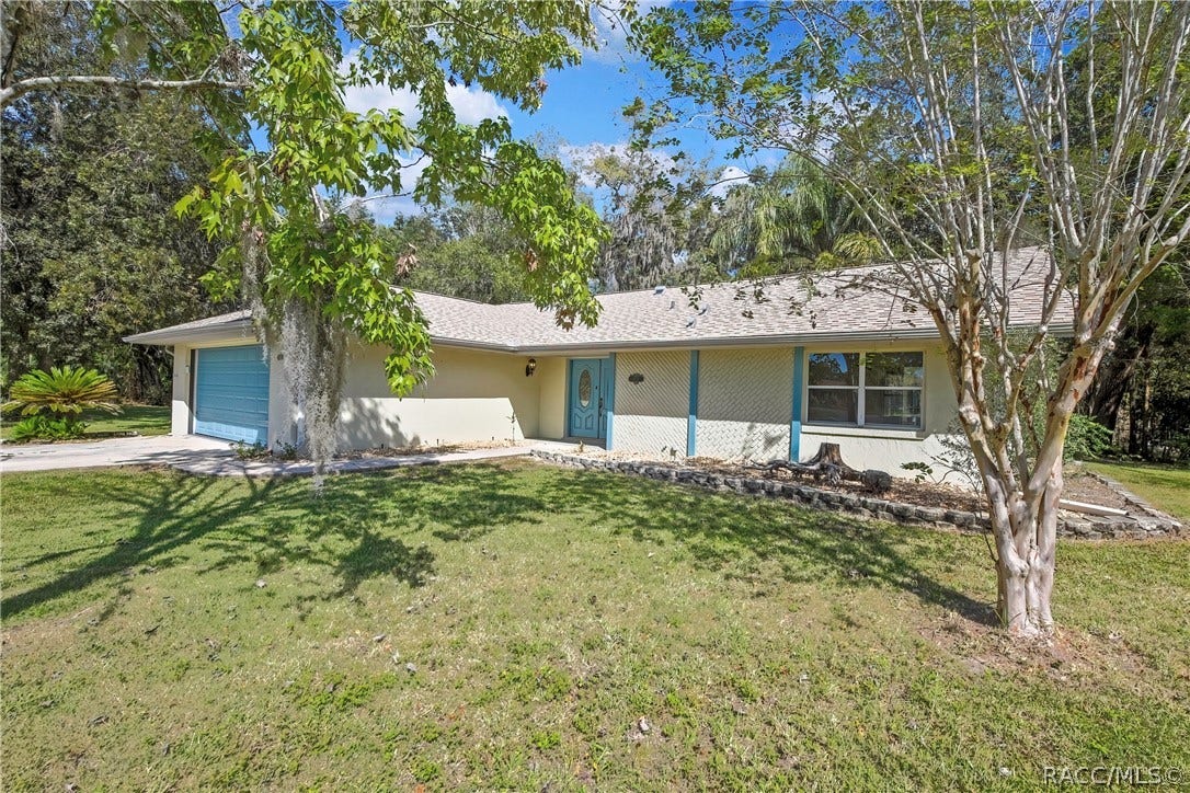 4737 S Sawmill Way, Homosassa, FL 34448 Front of the house seen from across the lawn, framed by tree branches with Spanish moss in the foreground: blue garage door at the left, blue entry door with oval glass in the centre, lattice wall section, a large window, a low stone retaining border along the front bed, a palm plant at the left, a multi-trunk tree at the right, green lawn, trees and palms behind the house, blue sky.