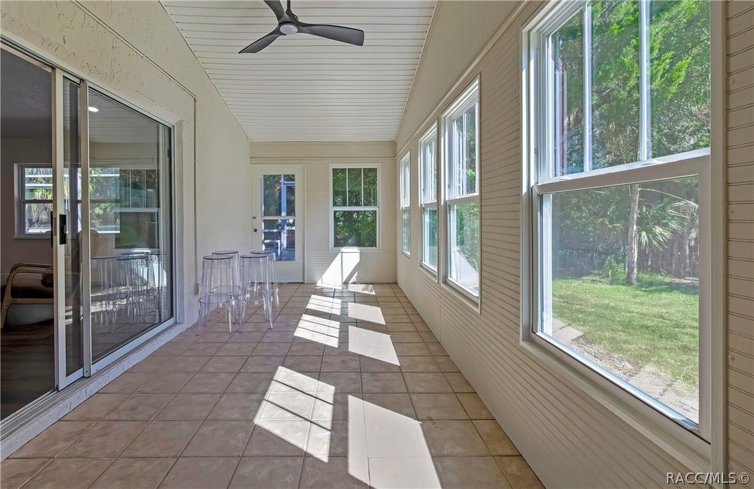 4737 S Sawmill Way, Homosassa, FL 34448 Screened porch, long view: tiled floor, white beadboard ceiling with a black ceiling fan, windows at the right looking onto the lawn and trees, sliding glass doors at the left to the living room, clear acrylic stools, and a white door and a window at the far end, with sunlight patches across the floor.