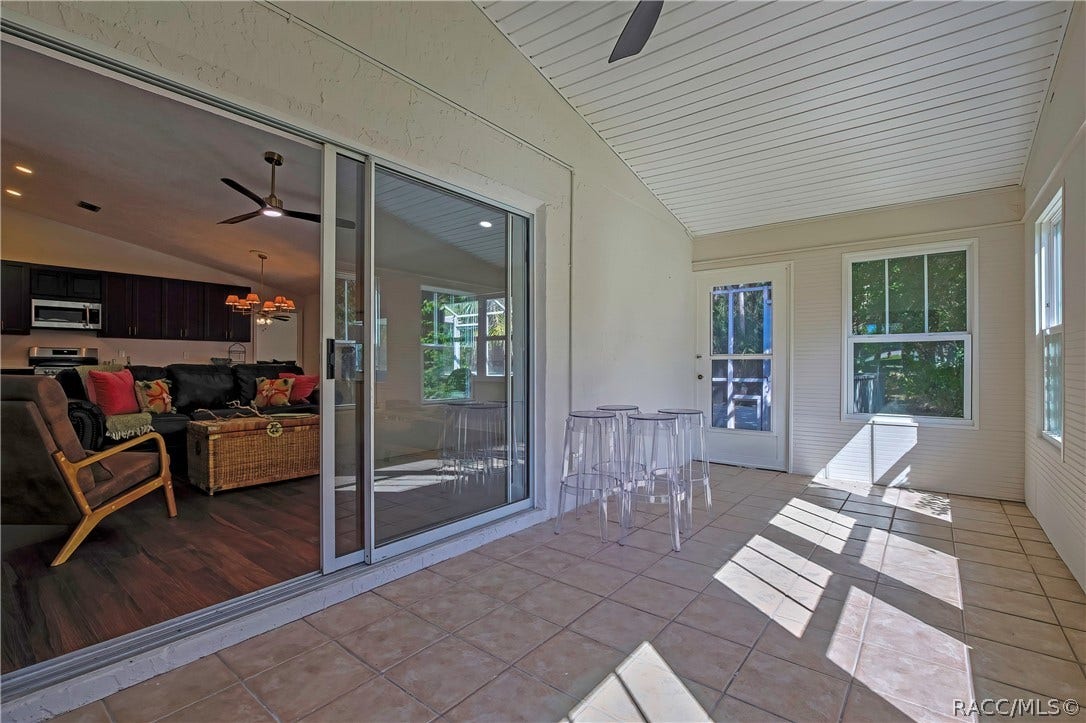 4737 S Sawmill Way, Homosassa, FL 34448 Screened porch from the other end: tiled floor, white beadboard ceiling, sliding glass doors at the left to the living room and kitchen, with the sofa, wicker trunk and armchair visible through them, clear acrylic stools in the centre, and a white door and a window at the far end, with sunlight patches across the floor.