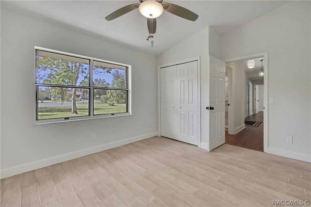 4737 S Sawmill Way, Homosassa, FL 34448 Empty bedroom: light plank tile floor, a large double window looking onto the street, front lawn, trees and neighbouring houses, white double closet doors in the centre, an open doorway at the right to a hallway with a dark wood floor and pendant lights, a ceiling fan with a dome light, and white walls. No furniture.