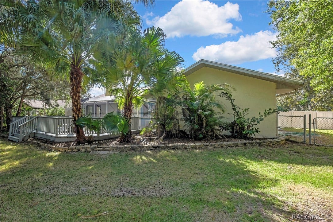4737 S Sawmill Way, Homosassa, FL 34448 Rear of the house from the lawn: light stucco walls, a shingle roof, a stucco chimney with a metal cap, a screen enclosure with blue doors, a grey wood deck with steps and railing, palm trees, a fenced lawn in the foreground, and neighbouring houses and chain-link and vinyl fences at the sides.