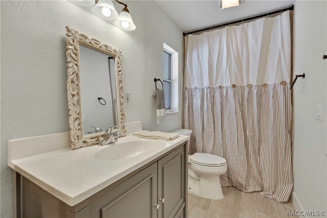 4737 S Sawmill Way, Homosassa, FL 34448 Primary bathroom, other angle: grey vanity with a white counter and an ornate white-framed mirror, a chrome faucet, a folded towel on the counter, a white toilet, a tub/shower with a white-and-striped curtain on a black rod, a narrow window, light plank tile floor, a 3-light fixture at the top of the frame, and a ceiling vent.