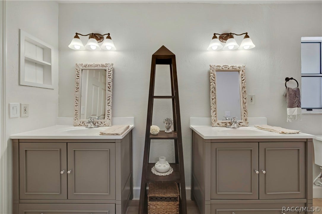 4737 S Sawmill Way, Homosassa, FL 34448 Primary bathroom with dual vanities: two grey vanity cabinets with white counters and chrome faucets, two ornate white-framed mirrors, a 3-light fixture above each mirror, a dark ladder shelf between the sinks with a bust, a white flower, a vase and a basket below, folded towels on each counter, a recessed wall niche at the left, a towel ring with a grey-and-white towel and a window at the right, and the edge of a toilet at the far right.