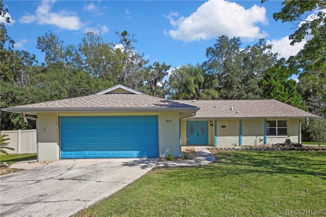 4737 S Sawmill Way, Homosassa, FL 34448 Straight-on front of the single-storey house: light cream stucco walls, grey shingle roof with a gable over the garage, wide blue panelled garage door, house number 4737 on the wall beside the garage, blue double entry doors with oval decorative glass, a wall light beside the doors, a lattice wall section and a large front window, concrete driveway and a curved concrete walkway to the entry, a stone-edged planting bed, green lawn, trees and palms, white vinyl fence at the left edge, blue sky