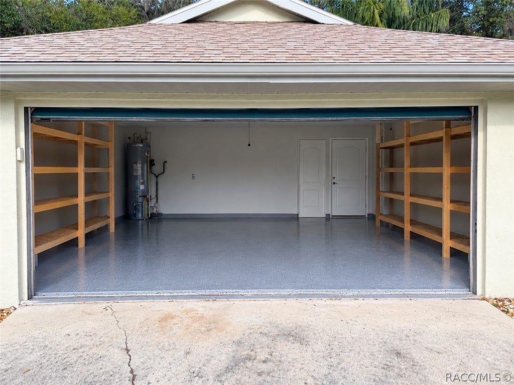 4737 S Sawmill Way, Homosassa, FL 34448 Clean, empty garage interior: smooth grey painted concrete floor, a white sectional garage door with metal rails and a garage door opener, a fluorescent tube light, a textured ceiling, beige walls, and a light wood empty shelving unit with two bays and four shelves against the back wall. No mower, boxes or clutter in this shot this is a different, newer garage photo than the two cluttered garage shots in the old set.