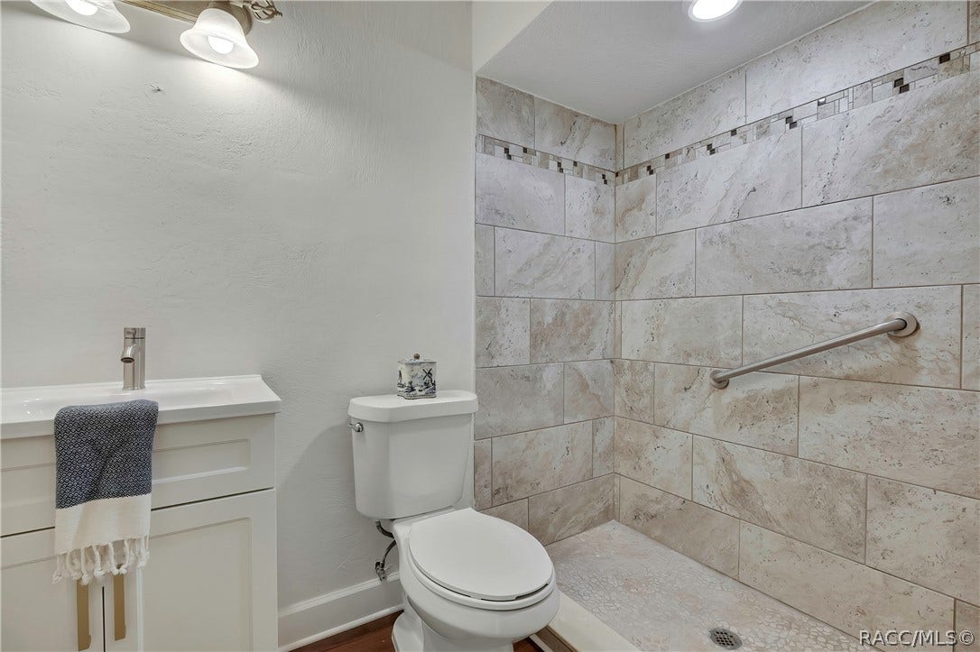 4737 S Sawmill Way, Homosassa, FL 34448 Bathroom: white vanity with a modern chrome faucet, a navy-and-white towel hanging over the vanity, a white toilet with a patterned container on the tank, a walk-in shower with stone-look tile walls, a mosaic accent strip, a grab bar and a pebble floor with a drain, recessed ceiling lights, and a 2-light fixture at the top left. This is the single-sink hall bath, not the primary bath.