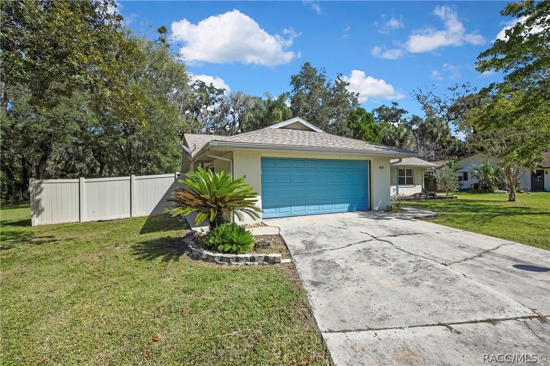 4737 S Sawmill Way, Homosassa, FL 34448 Front-garage angle: blue panelled garage door, house number 4737 on the wall beside it, wide concrete driveway in the foreground, white vinyl fence at the left, a palm plant in a stone-edged bed in front of the garage, green lawn, a neighbouring house at the right, trees, blue sky with white clouds.