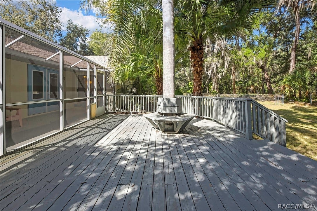 4737 S Sawmill Way, Homosassa, FL 34448 Wood deck: grey wood deck planks, railing, a built-in bench around a palm tree trunk, steps down to a fenced lawn, palm trees, oaks with Spanish moss, and a chain-link fence in the background.