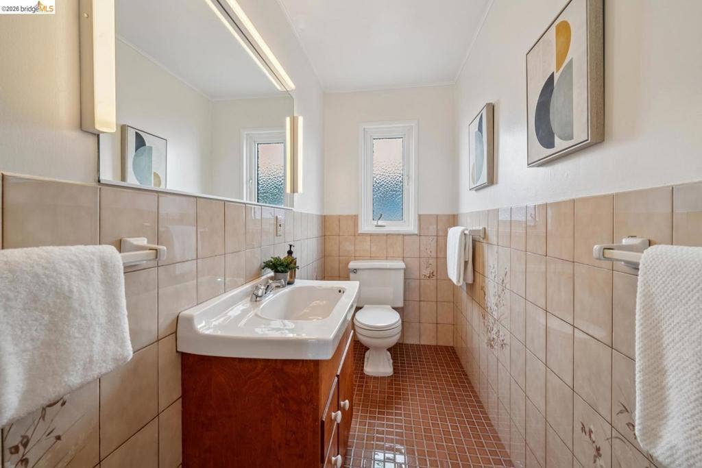 White ceramic toilet, window with textured glass, wood-finish vanity with an integrated sink, wall-mounted mirror, and ceramic wall tiling
