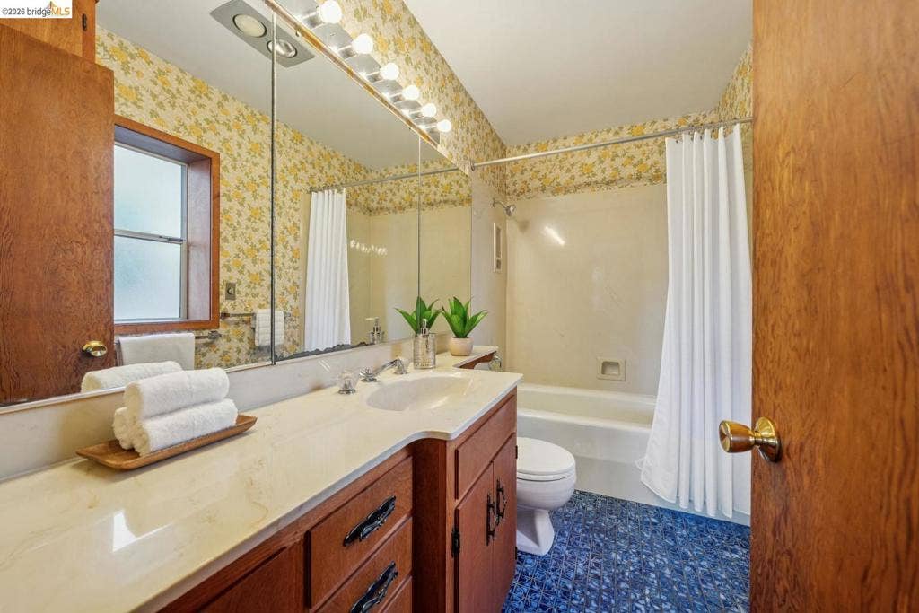Built-in vanity with an integrated sink and light-colored countertop, large wall-mounted mirror, overhead vanity lighting, window with frosted glass, and a bathtub with a shower curtain