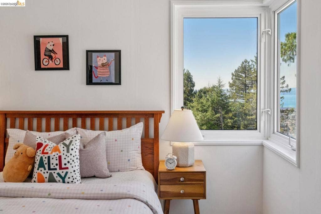 White wall finishes, natural wood headboard, corner window configuration, framed wall art, and a wood-finish nightstand