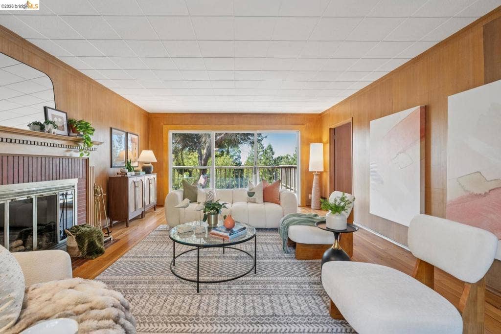 Wood paneling throughout, hardwood flooring, fireplace with a decorative mantel, expansive window with outdoor scenery, and a drop ceiling