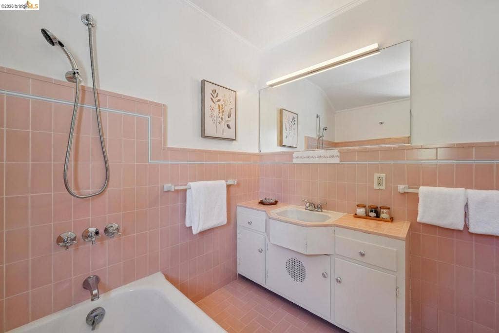 Bathroom featuring pink tiled walls with a light blue accent border, a white bathtub with a handheld shower, a white vanity with integrated sink, a large wall-mounted mirror, and overhead lighting