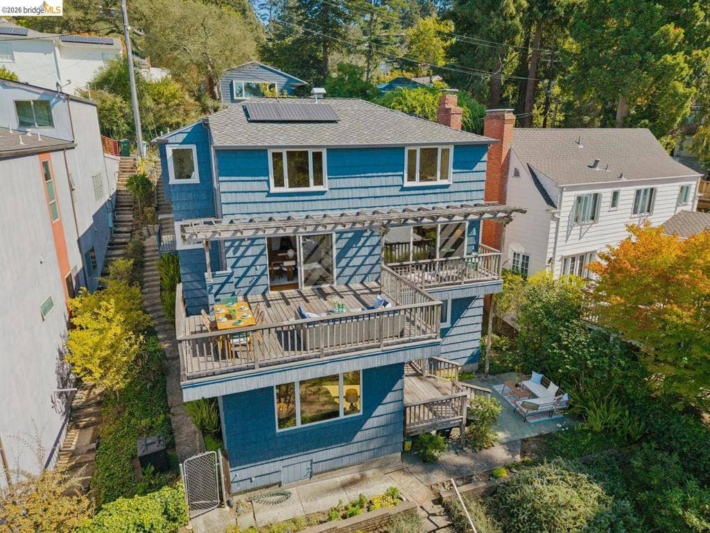 Multi-level exterior with blue siding, a multi-tiered deck with wood railing, a rooftop solar panel array, and an integrated pergola