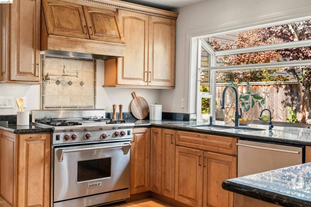 Wood cabinetry with upper crown molding, stainless steel range with pot filler, granite countertops, tile backsplash, and a large garden window