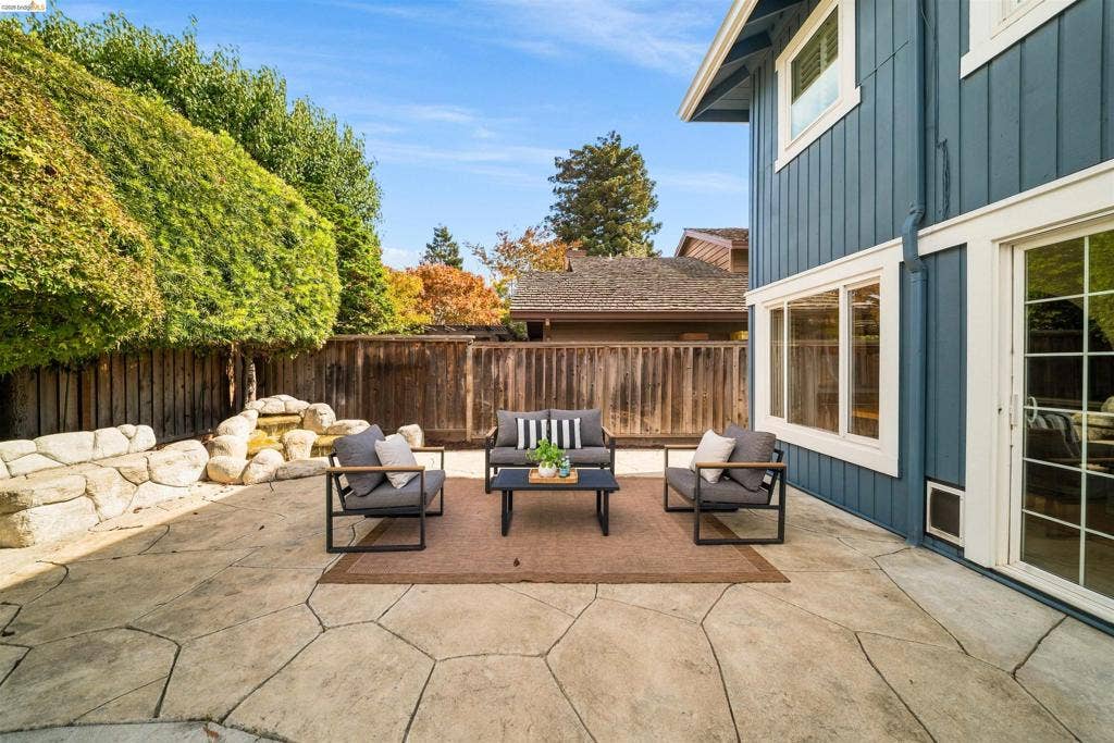 Expansive stamped concrete patio with rock water feature, bordered by a wooden fence with mature foliage, and featuring sliding glass doors and large windows on the blue siding exterior
