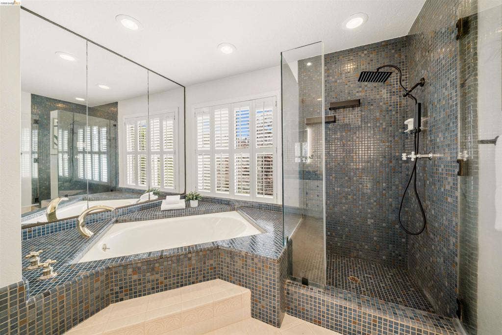 Corner soaking tub with mosaic tile surround, walk-in shower with mosaic tile, glass enclosure, and matte black fixtures, and windows with plantation shutters