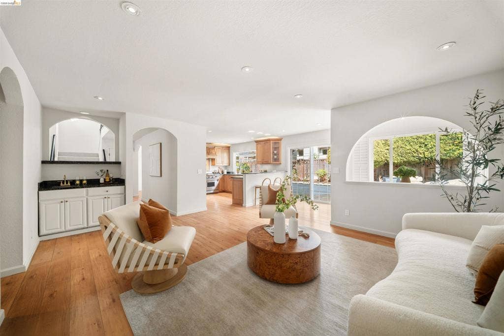 Recessed lighting, wood-finish flooring, arched doorways, built-in wet bar with white cabinetry and dark countertop, and a window with garden scenery