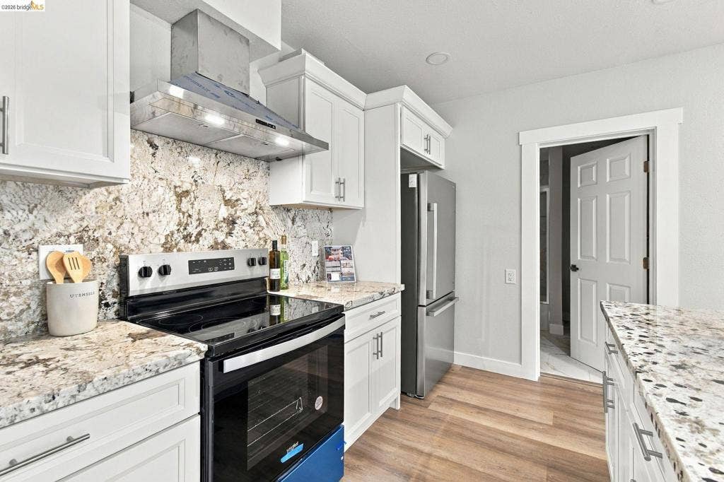 Kitchen featuring white cabinetry, stainless steel appliances, a range hood, wood-finish flooring, and stone countertops
