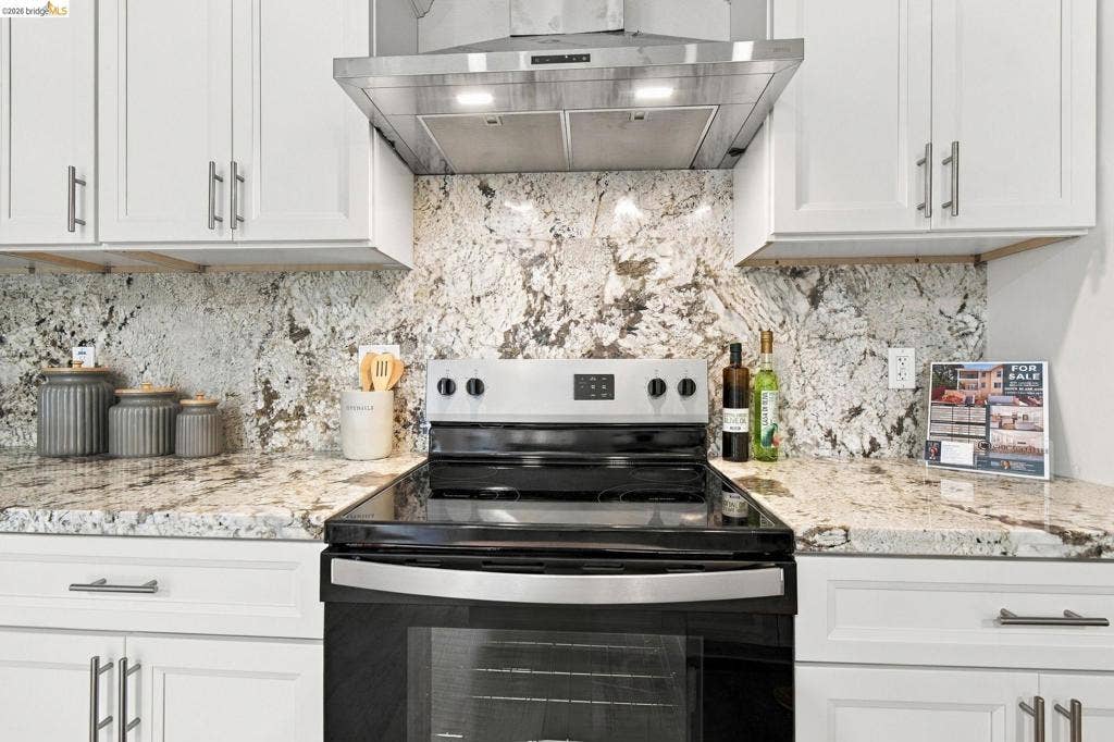 White cabinetry with brushed nickel hardware, expansive granite countertops, full-height granite backsplash, stainless steel range hood, and a black electric range