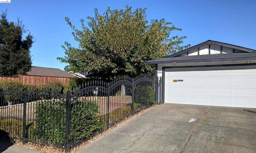 145 Viking Way, Pittsburg, CA 94565 White panel garage door, concrete driveway, black wrought iron fence with dual gates, low-maintenance landscaping, and mature tree