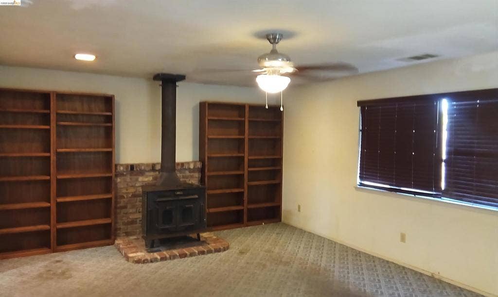 145 Viking Way, Pittsburg, CA 94565 Wood-burning stove with a brick hearth and surround, flanked by built-in wood shelving, ceiling fan with integrated lighting, and a large window with blinds
