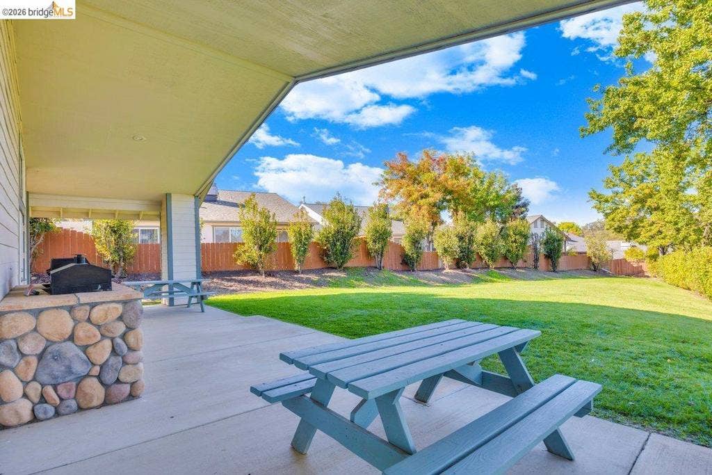 981 Country Ln, Angels Camp, CA 95222 Covered patio with a stone-clad built-in barbecue, concrete surface, and expansive grassy yard featuring mature trees and a wood fence