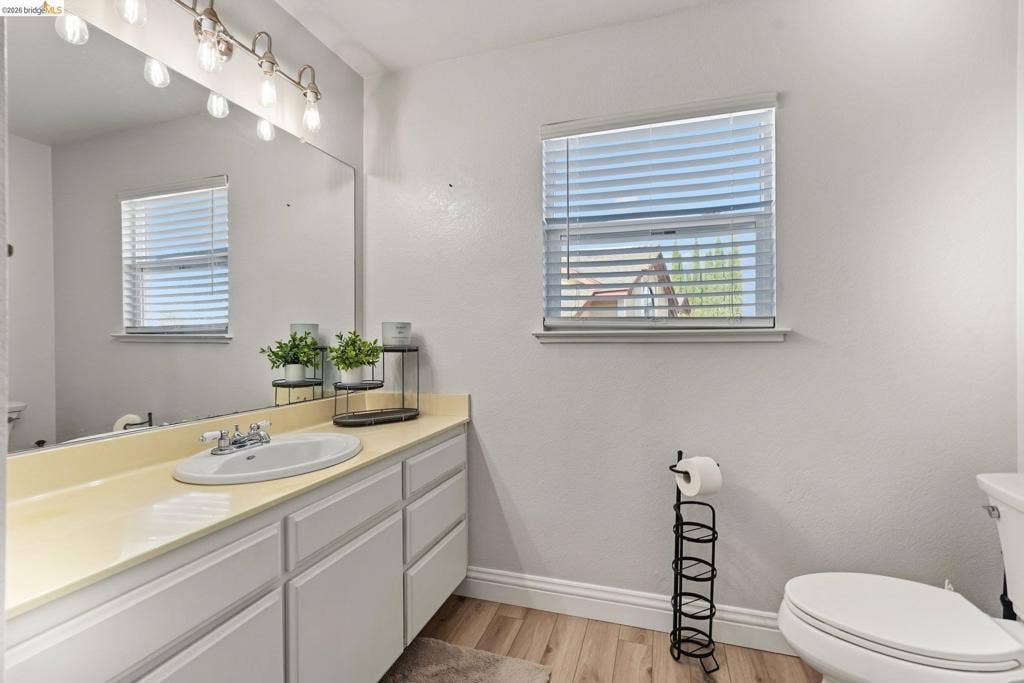 304 Greystone Dr, Antioch, CA 94509 White vanity cabinetry with a solid-surface countertop, an integrated oval basin, a full-width wall mirror, wood-finish flooring, and horizontal window blinds