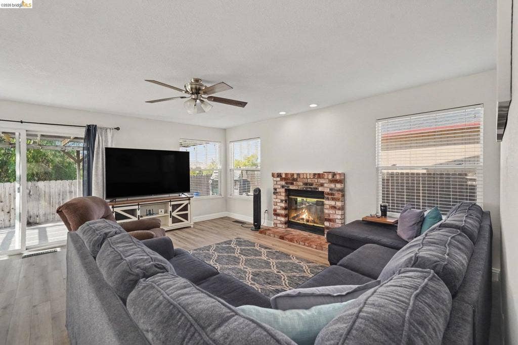 304 Greystone Dr, Antioch, CA 94509 Living area featuring wood-finish flooring, a brick fireplace with a glass-front insert, multiple windows, a ceiling fan, and sliding glass patio doors